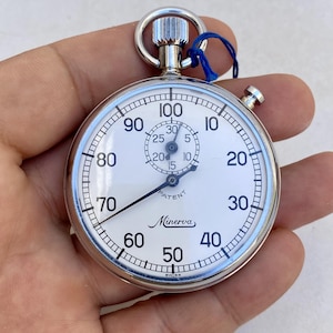 Può includere: Un cronometro Minerva argentato con quadrante bianco e numeri neri. L'orologio presenta un piccolo quadrante dei secondi e una lancetta dei secondi grande. La parola "Minerva" è stampata sul quadrante. Un cordino blu è attaccato in alto.