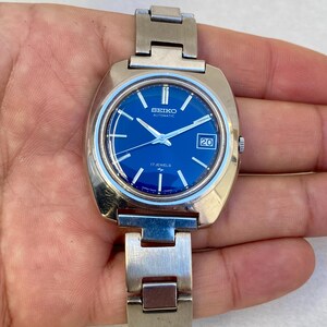 Puede incluir: Un reloj automático Seiko vintage con caja rectangular de acero inoxidable y esfera azul vibrante. El reloj presenta una ventana de fecha a las 3 en punto y un brazalete plateado. La esfera dice "SEIKO AUTOMATIC 17 JEWELS".