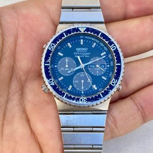 May include: A silver Seiko Quartz Chronograph Sports 100 watch with a blue dial and bezel. The watch features multiple sub-dials, a tachymeter scale, and the date. The watch band is made of silver metal links.