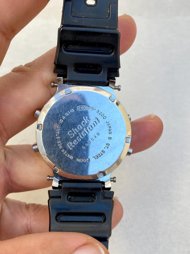 May include: A black Casio digital watch with a silver-tone back. The watch has a black strap and the text "Shock Resistant" and "Water Resistant 200M". The model number is 240DW-5200.
