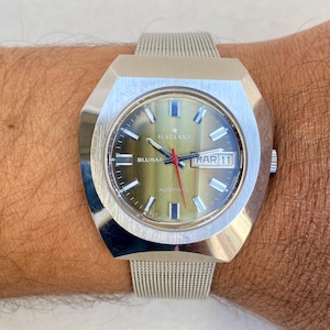 May include: A silver-toned wristwatch with a green face and a mesh band. The watch face displays the date as "MAR 11" and the brand name "RADIANT" above the hands. The watch is on a wrist.