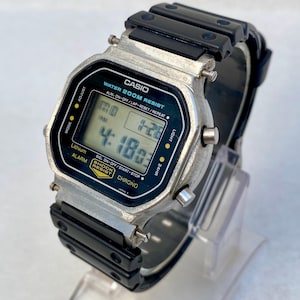 May include: A Casio digital watch with a black resin strap and a silver-tone case. The watch face shows the time and other functions. The watch is labelled "WATER 200M RESIST" and "SHOCK RESIST".