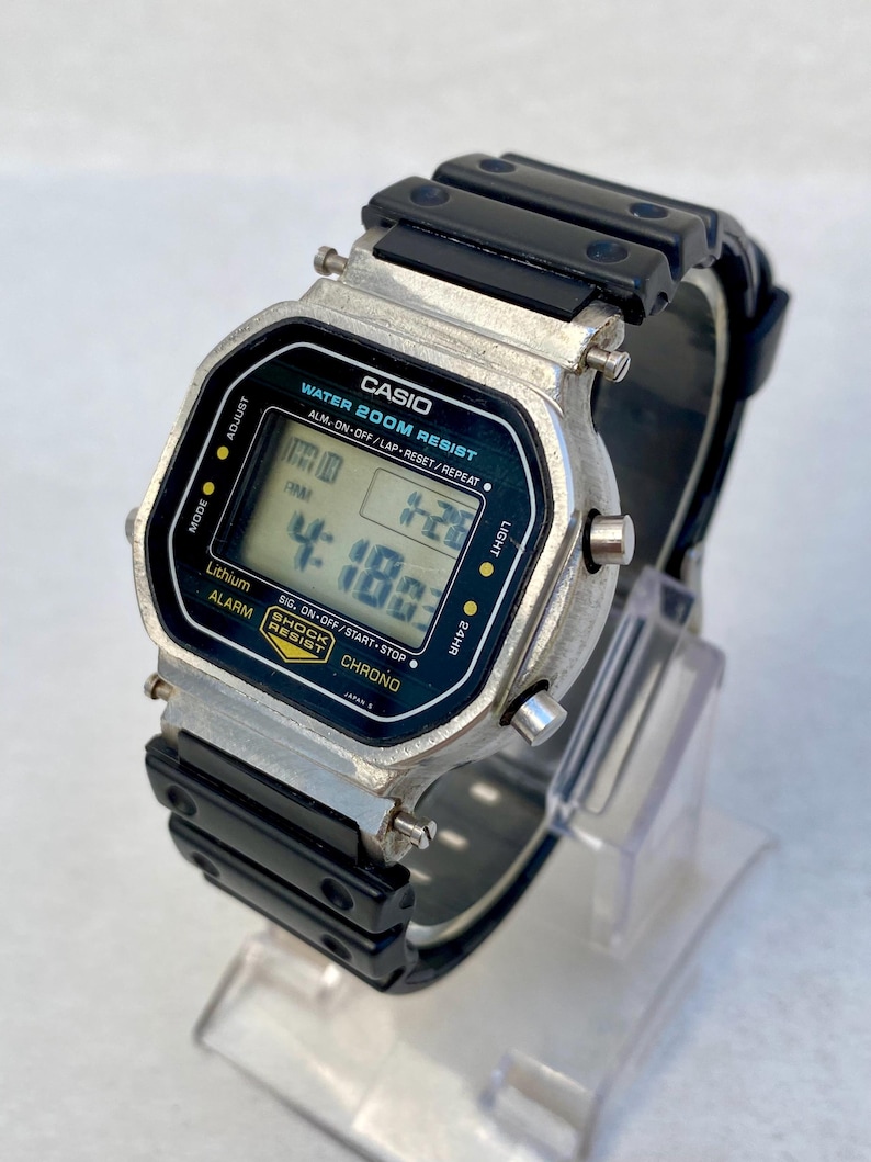May include: A Casio digital watch with a black resin strap and a silver-tone case. The watch face shows the time and other functions. The watch is labelled "WATER 200M RESIST" and "SHOCK RESIST".