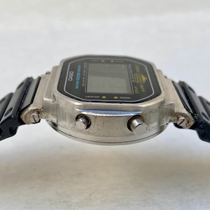 May include: A silver Casio digital watch with a black rectangular face and black strap. The watch face displays the Casio logo and other text. The watch has two buttons on the side and a black, segmented strap.