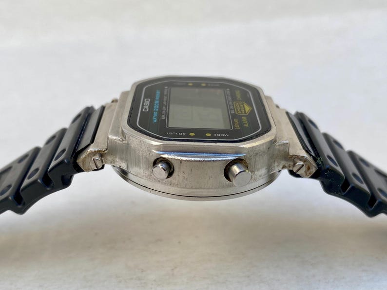 May include: A silver Casio digital watch with a black rectangular face and black strap. The watch face displays the Casio logo and other text. The watch has two buttons on the side and a black, segmented strap.