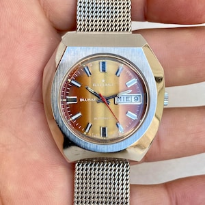 May include: A vintage Radiant Bluemar automatic wristwatch with a silver-tone case and mesh band. The watch face has a gradient brown and red color scheme with the day and date displayed. The watch is held by a hand.