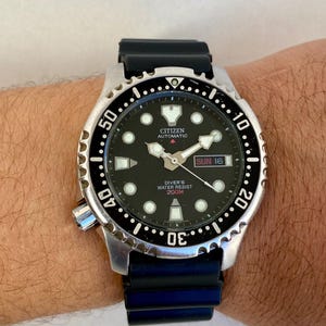 Peut inclure: Une montre de plongée automatique Citizen noire avec un bracelet en caoutchouc noir. Le cadran de la montre a un fond noir avec des marques blanches et une fenêtre de date qui indique "SUN 16". La lunette est noire avec des marques blanches et indique "DIVER'S WATER RESIST 200M".