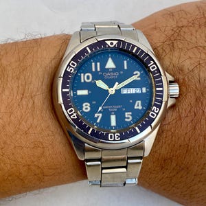 Peut inclure: Une montre Casio quartz argentée avec un cadran bleu et une lunette noire. La montre a un guichet date qui affiche "FRI 27" et une étanchéité de 100m.