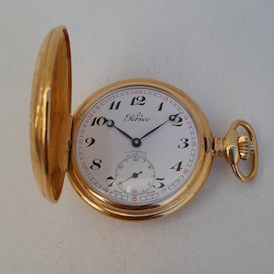 Puede incluir: Un reloj de bolsillo dorado con una esfera blanca y agujas negras. El reloj tiene un movimiento Incabloc de 17 joyas y está hecho en Suiza. La esfera del reloj tiene el texto "Perseo" y los números del 1 al 12 alrededor del borde.