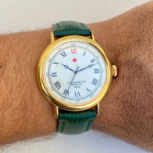 Puede incluir: Un reloj de pulsera dorado con esfera blanca, números romanos negros y una cruz roja. El reloj tiene una correa de cuero verde y el texto "CHRONOMETER MEDICAL 1910".