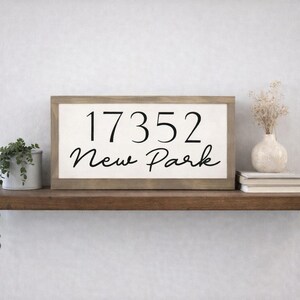 May include: A rectangular wooden sign with the numbers "17352" and the words "New Park" in black script. The sign is displayed on a dark brown shelf, alongside a small white vase, books, and a potted plant.