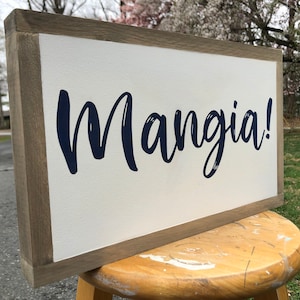 May include: A rectangular wooden sign with a white background and a dark blue script that reads "Mangia!". The sign has a light brown wooden frame.