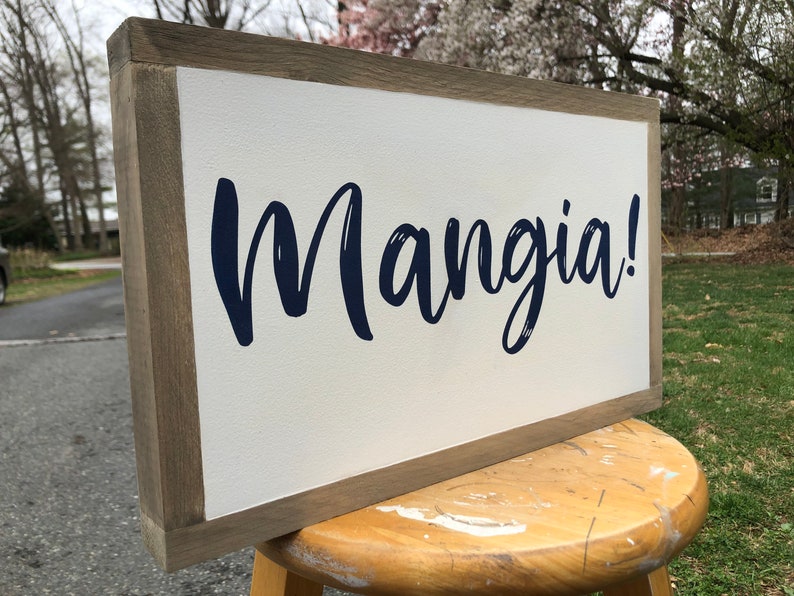 May include: A rectangular wooden sign with a white background and a dark blue script that reads "Mangia!". The sign has a light brown wooden frame.