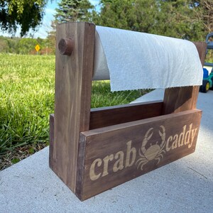 May include: A wooden crab caddy with a roll of paper towels. The caddy is stained a medium brown and features the words "crab caddy" and a crab graphic. The caddy is outdoors on a concrete surface.