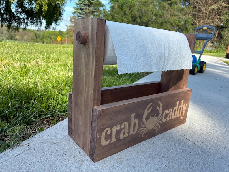 May include: A wooden crab caddy with a roll of paper towels. The caddy is stained a medium brown and features the words "crab caddy" and a crab graphic. The caddy is outdoors on a concrete surface.