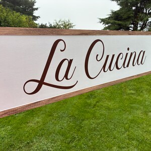 Puede incluir: Un letrero rectangular de madera con fondo blanco y un marco de madera te&ntilde;ida de color marr&oacute;n oscuro. El texto La Cucina est&aacute; impreso en una elegante letra cursiva de color marr&oacute;n oscuro en el centro de la superficie blanca del letrero.