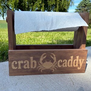 May include: A wooden paper towel holder with a rectangular base and a roll of white paper towels. The front of the holder is engraved with the words "crab caddy" and a crab illustration. The holder is made of dark brown wood.