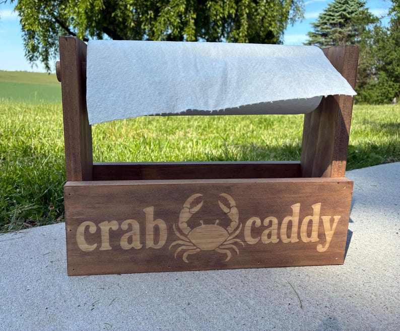 May include: A wooden paper towel holder with a rectangular base and a roll of white paper towels. The front of the holder is engraved with the words "crab caddy" and a crab illustration. The holder is made of dark brown wood.
