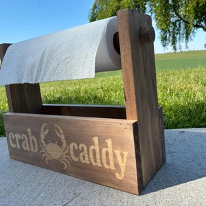 May include: A wooden crab caddy with a roll of paper towels. The caddy is made of stained wood and features the words "crab caddy" and a crab illustration. The caddy is designed to hold paper towels and is suitable for outdoor use.