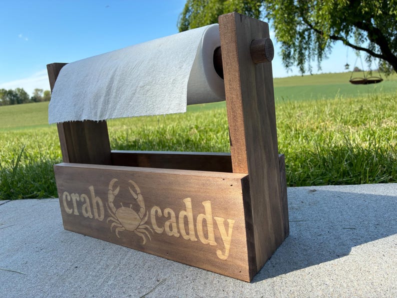 May include: A wooden crab caddy with a roll of paper towels. The caddy is made of stained wood and features the words "crab caddy" and a crab illustration. The caddy is designed to hold paper towels and is suitable for outdoor use.