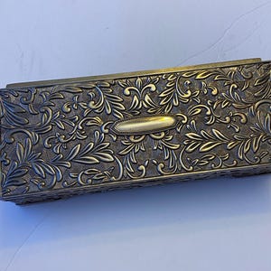 Op de afbeelding: Een rechthoekige metalen doos met een antieke messing afwerking, voorzien van een ingewikkeld reliëfpatroon van bloemen en krullen op het deksel. Een glad, leeg, langwerpig ovaal plaatje is gecentreerd op de bovenkant van het deksel.