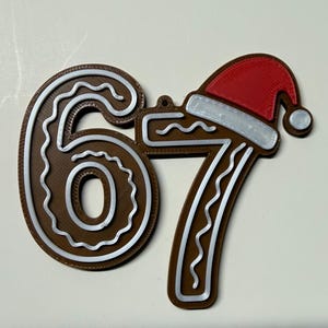 May include: A brown Christmas ornament in the shape of the numbers 67. The number 7 is topped with a red Santa hat with a white trim. The numbers have a white, piped frosting design.