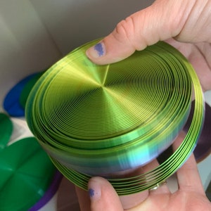 May include: A green iridescent ribbon coiled into a spiral shape. The ribbon is made of a thin, flexible material and has a metallic sheen.