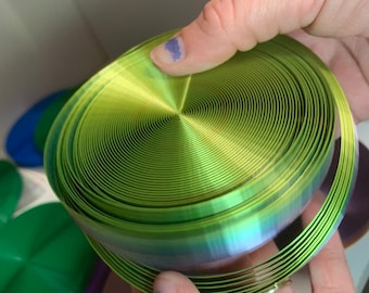 May include: A green iridescent ribbon coiled into a spiral shape. The ribbon is made of a thin, flexible material and has a metallic sheen.