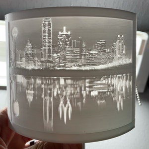 May include: A white 3D printed lampshade with a cityscape design. The cityscape is of a city skyline with tall buildings and a river. The cityscape is reflected in the water below.