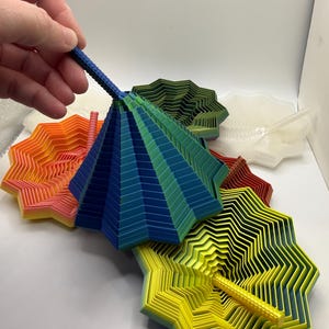 Könnte beinhalten: Eine Sammlung farbenfroher, geschichteter Papierkunstwerke in verschiedenen Formen und Größen. Die Stücke umfassen eine blau-grüne schirmartige Struktur und sternförmige Designs in Gelb, Orange und Grün. Die Kunstwerke werden auf einer weißen Oberfläche präsentiert.