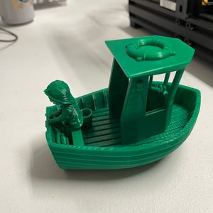 May include: A green plastic toy boat with a small figure of a person sitting in the front. The boat has a life preserver on the top and a wooden deck.