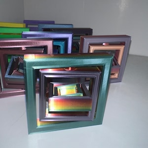 May include: A set of nested square frames made of 3D printed plastic. The frames are in various colors, including green, purple, brown, and gold. The frames are arranged in a stack, with the largest frame at the bottom and the smallest frame at the top.