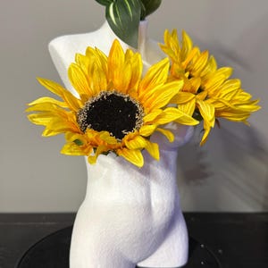 May include: A white bust-shaped vase holds bright yellow sunflowers with dark centers and green foliage. The vase is a stylized representation of a torso, with a textured surface. The sunflowers add a pop of color against the neutral background.