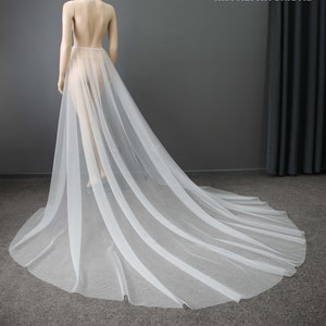 May include: A sheer white bridal skirt with a long train, draped on a mannequin. The skirt is made of a lightweight, flowing fabric, with vertical stripes. The mannequin is positioned against a gray backdrop.