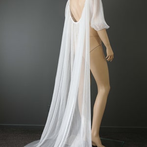 May include: A white, pleated bridal cape with a low back and short sleeves, displayed on a mannequin. The cape flows down to the floor, creating a dramatic effect. The brand name "KIM ALPHA BRIDAL" is visible in the upper left corner.