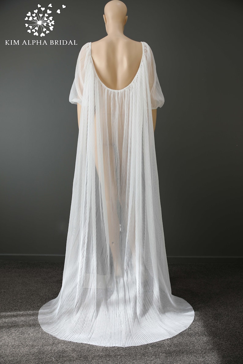 May include: A white, pleated bridal cape with a flowing design. The cape features a scoop back and short sleeves. The Kim Alpha Bridal logo is visible in the upper left corner.