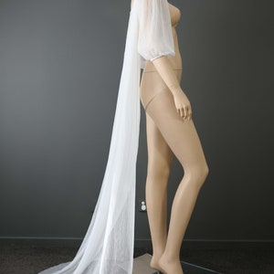 May include: A white, pleated bridal sleeve and cape, draped on a mannequin. The sleeve has a puffed shoulder and a long, flowing cape. The mannequin is on a glass base. The text "KIM ALPHA BRIDAL" is in the upper left corner.