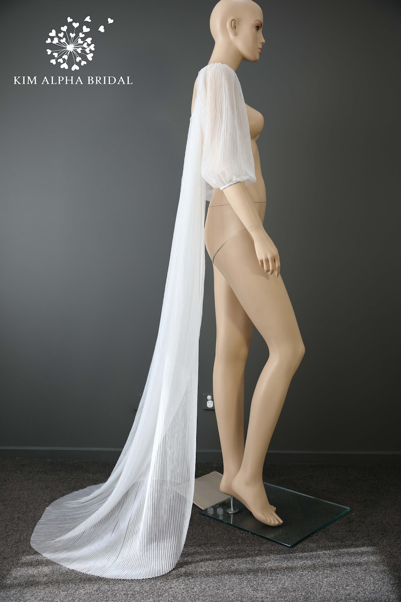 May include: A white, pleated bridal sleeve and cape, draped on a mannequin. The sleeve has a puffed shoulder and a long, flowing cape. The mannequin is on a glass base. The text "KIM ALPHA BRIDAL" is in the upper left corner.