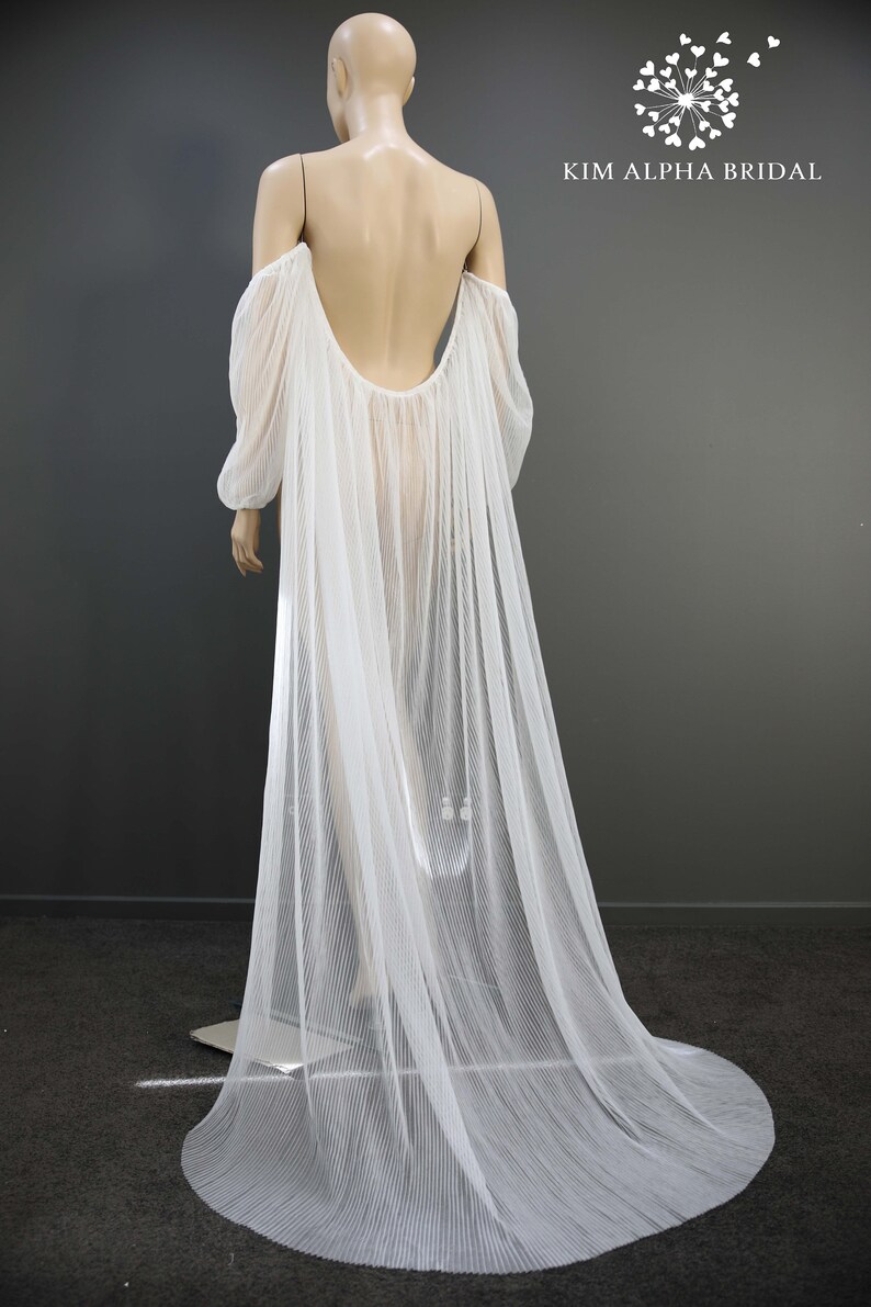 May include: A sheer white bridal cape with long sleeves and a flowing train. The cape is draped over a mannequin, showcasing its delicate, pleated fabric and open back design. The Kim Alpha Bridal logo is visible.