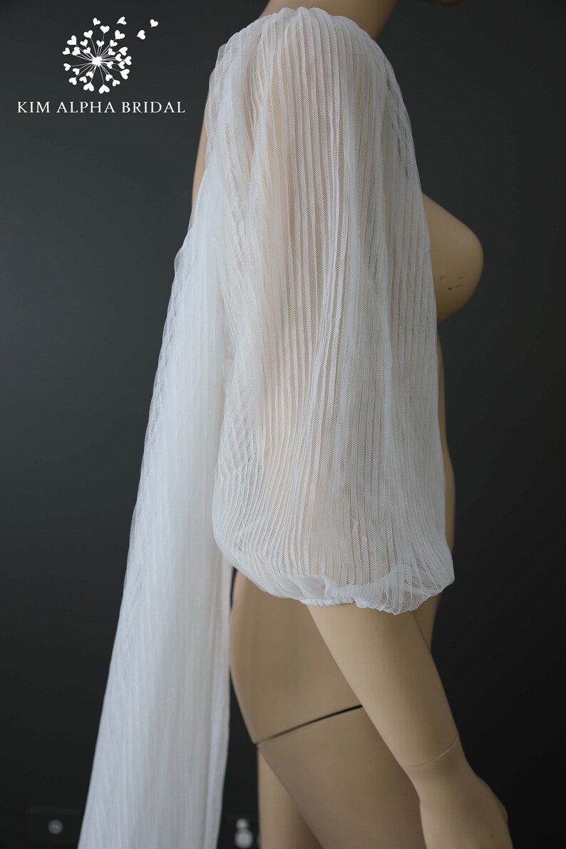 May include: White, pleated bridal sleeve with a long, flowing panel. The sleeve is made of a sheer, textured fabric. The Kim Alpha Bridal logo is visible in the upper left corner. The sleeve is displayed on a mannequin.