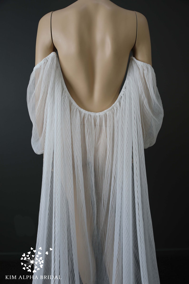 May include: A white, off-the-shoulder bridal cape with a low back. The cape is made of a pleated, sheer fabric that drapes over the shoulders and back. The cape is displayed on a mannequin against a dark gray background. The brand name "KIM ALPHA BRIDAL" is visible.