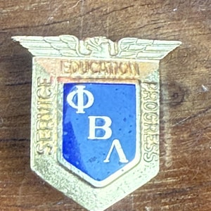 Puede incluir: Un pin dorado con un centro azul, con las letras griegas Phi, Beta y Lambda. El pin tiene forma de escudo, con las palabras "SERVICE", "EDUCATION" y "PROGRESS" inscritas en los bordes.