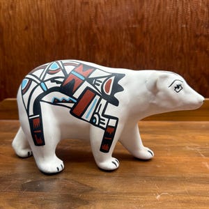 Puede incluir: Una figura de oso polar de cerámica blanca con intrincados patrones geométricos en negro, rojo y azul claro en el lomo y los costados. El oso se muestra en posición de pie con un acabado mate suave y rasgos faciales detallados.