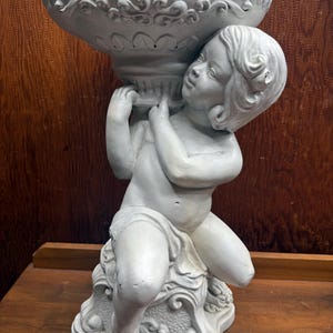Könnte beinhalten: Graue Cherub-Statue, die eine große, verzierte Schale hält. Der Cherub kniet auf einem dekorativen Sockel. Die Schale hat einen detaillierten, gewellten Rand und wirbelnde Muster. Die Statue besteht aus einem hellen Material, möglicherweise Beton oder Gips.