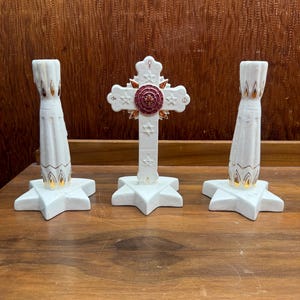 May include: A set of three white ceramic religious items. The set includes two candle holders and a cross. The candle holders have a star-shaped base and gold accents. The cross has a circular emblem in the center.