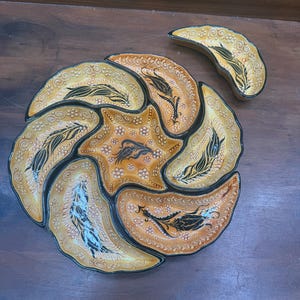 Könnte beinhalten: Ein dekoratives Servierschalen-Set mit einer sternförmigen Zentralschale und sechs umliegenden blütenblattförmigen Fächern. Die Keramikschale hat eine warme Farbpalette aus Gold, Orange und Schwarz mit aufwendigen Blumen- und Blattmustern. Eine passende halbmondförmige Schale ist ebenfalls enthalten.