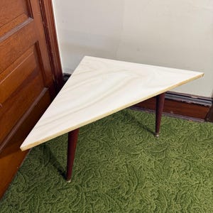 Puede incluir: Una pequeña mesa triangular de madera con una tapa de color claro y tres patas rojas oscuras. Las patas miden unos 30 cm de largo. La mesa está sobre una alfombra verde estampada, con una puerta de madera y una pared blanca al fondo.