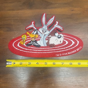 May include: A red and white patch featuring Bugs Bunny, Daffy Duck, Taz, and Tweety Bird. The patch is oval-shaped with a white spiral design and the text "TM © 1994 WARNER BROS." The patch measures approximately 5 inches in length.