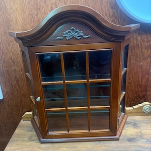 Pode incluir: Uma vitrine de madeira com porta de vidro e prateleiras. O armário tem um topo curvo com detalhes decorativos e um acabamento castanho escuro. A porta de vidro é dividida em vários painéis e o armário está sobre uma superfície de madeira.