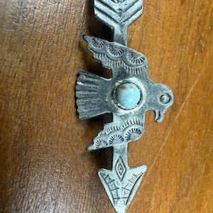 Puede incluir: Colgante de metal plateado con diseño de flecha, figura aviar y piedra turquesa. El colgante presenta intrincados detalles grabados y una estética vintage, adecuado para joyería o proyectos de artesanía.